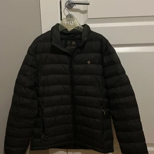 Svart pufferjacka från Morris - Säljer en stilren olivgrön (i ljuset ser den svart ut) pufferjacka från Morris. Jackan har en diskret broderad detalj på bröstet och dragkedja framtill. Perfekt för kyligare dagar. En klassisk och tidlös modell för alla tillfällen. Hör av er vid fler frågor och bilder! Mvh