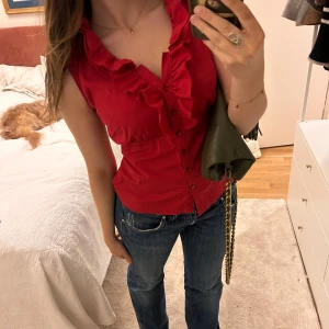 Röd blus med volangdetaljer - Säljer en snygg röd blus med volangdetaljer och knappar framtill. Blusen har en figurnära passform och är ärmlös, vilket gör den perfekt för varmare dagar. Den är både stilren och trendig, och passar bra till jeans eller kjol.