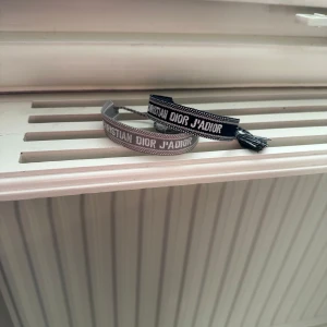 Armband från Christian Dior - Två stilrena armband från Christian Dior med texten 'J'ADIOR'. Ett armband är i grått och det andra i svart, båda med vita detaljer. Perfekta för att ge en elegant touch till din stil.