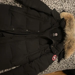 Svart dunjacka från Canada Goose - Säljer en svart dunjacka från Canada Goose med pälskantad huva. Jackan har en klassisk design med dragkedja och knappar framtill. Perfekt för kalla vinterdagar. Märkeslogga på ärmen ger en exklusiv touch.