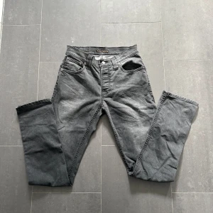 Grå jeans från Nudie Jeans Grim Tim - Säljer ett par grå jeans från Nudie Jeans, modellen Grim Tim. Jeansen har fem fickor, raka ben och klassisk knappgylf. Tillverkade i bomull med en snygg tvättad grå färg och diskreta sömmar.