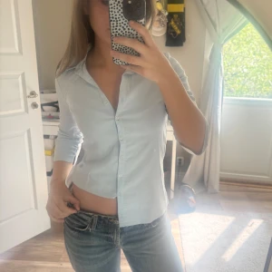 Ljusblå skjorta med knappar - Säljer en ljusblå skjorta med klassisk krage och långa ärmar. Skjortan har en figurnära passform och stängs med knappar framtill. Perfekt att styla med jeans för en clean look.