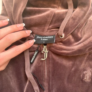 Brun velour hoodie från Juicy Couture - Mysig brun hoodie i velour från Juicy Couture med dragkedja framtill och huva. Jackan har långa ärmar, fickor på sidorna och broderad logga på bröstet.  Bra skick! Ej använd så jätte mycket! 