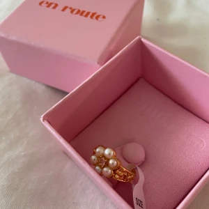 Guldring med pärlor från en route - Aldrig använd! Säljer en vacker guldfärgad ring från en route med dekorativa vita pärlor och små gnistrande stenar. Ringen har en unik design där pärlorna bildar ett blommönster. Snyggt att matcha med andra chunky ringar!