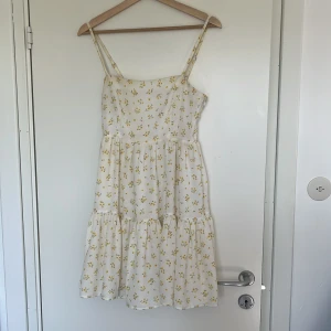 Vit blommig klänning från Lisa & Lena for NA-KD - Supersöt vit klänning med små gula blommor från Lisa & Lena for NA-KD. Axelbandslös modell med volangdetaljer och luftig passform. Perfekt för varma dagar och somriga tillfällen.