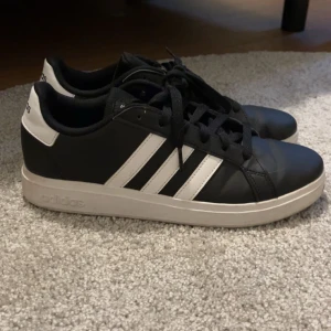 Svarta och vita sneakers från Adidas - Säljer ett par klassiska svarta sneakers från Adidas med vita ränder och vit sula. Skorna har snörning och är i syntetmaterial. Perfekta för dig som gillar en stilren och sportig look.