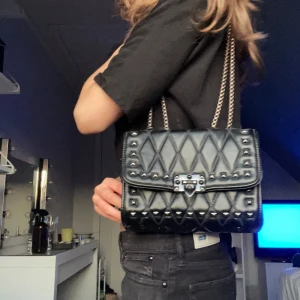 Svart quiltad axelväska med kedja - Säljer en svart quiltad axelväska med silverfärgad kedja och nitar. Väskan har ett lock med spänne framtill och ett elegant rutmönster. Perfekt för att lyfta din outfit med en edgy touch.