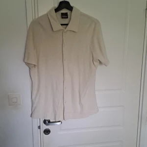 Beige kortärmad frottéskjorta från Oscar Jacobson - Säljer en stilren kortärmad frottéskjorta från Oscar Jacobson. Skjortan har knäppning framtill och är tillverkad i ett mjukt frottématerial. Perfekt för en avslappnad och trendig look.