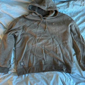 Grå hoodie från Morris - Säljer en grå hoodie från Morris med dragsko i huvan och en broderad logga på bröstet. Tröjan har en mjuk insida och ribbade muddar vid ärmslut och nederkant. Storlek XL men passar M skulle jag säga. Säljer eftersom den är för liten för mig. Aldrig använd. 
