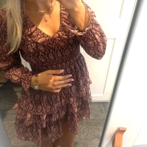 volangklänning  - Kort mönstrad klänningv-ringad och långa ärmar. Smockad midja och volangdetaljer på kjolen. Perfekt för dig som gillar romantisk och bohemisk stil.