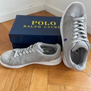 Grå sneakers från Polo Ralph Lauren - Snygga grå sneakers från Polo Ralph Lauren med vit sula och klassisk blå logga på sidan. Skorna har rund tå, platt sula och är tillverkade i mocka med snörning framtill. Perfekta för en stilren och avslappnad look. Skorna är också i nästan ny skick