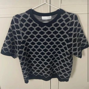 Svart stickad tröja med vitt mönster - Säljer en svart stickad topp med korta ärmar och vitt geometriskt mönster. Tröjan har rund halsringning och ribbade muddar vid ärmslut och nederkant. Perfekt för dig som gillar stilrena men ändå unika plagg.