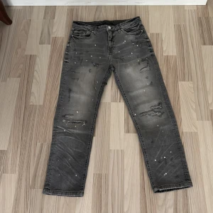 Grå jeans med slitningar från Supply & Demand - Säljer ett par grå jeans från Supply & Demand med coola slitningar och färgstänk. Byxorna har fem fickor, raka ben och klassisk knappgylf. Perfekta för dig som gillar en streetwear-look.