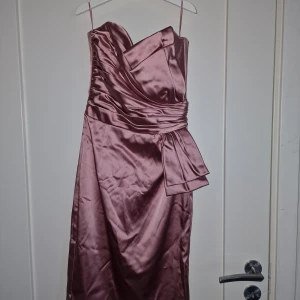 Rosa Klänning - Elegant klänning från Belle by OASIS i storlek 8/34. Stilren design i glansigt roséguld satin, perfekt för fest, bröllop eller bal. Figurnära passform med vackert draperat liv och dekorativ rosett i midjan. Justerbara axelband och dold dragkedja baktill. Klänningen är i mycket fint skick och sitter otroligt smickrande på kroppen. 📦Snabb leverans - Skickas samma dag📦 Kontakta mig gärna vid frågor eller intresse.
