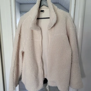 Beige teddyjacka med dragkedja - Mysig beige teddyjacka med hög krage och hel dragkedja framtill. Jackan har lång ärm och är tillverkad i ett fluffigt fleecematerial. Perfekt för kyliga dagar och enkel att matcha med olika outfits.
