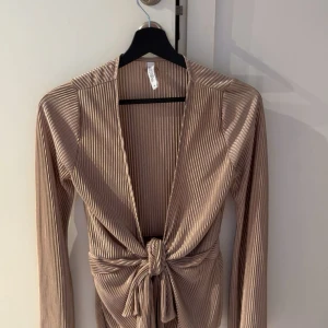 Beige ribbad knytblus från H&M - Säljer en beige ribbad blus från Bubbleroom med lång ärm och djup v-ringning. Toppen har knyt framtill i midjan och är i ett mjukt, stretchigt material. Perfekt för att lyfta en outfit med sin snygga passform och trendiga look. Storlek M💕