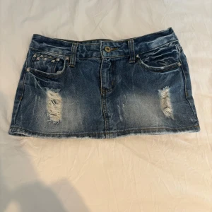 Jeanskjol med nitar från Sinmau - Kort jeanskjol i blå denim med slitna detaljer och silvriga nitar på fickorna fram och bak. Midjemått: 38 cm rakt över, kjollängd: 27 cm