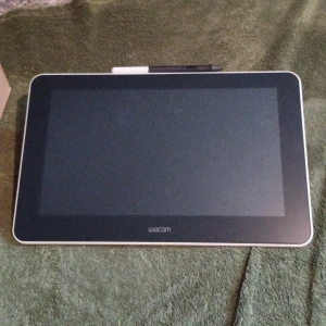 Wacom ritplatta med penna - Digital ritplatta från Wacom med stor svart skärm och vit ram. Medföljer en svart digital penna och laddare med tillhörande kablar. Perfekt för digitalt skapande och illustration.