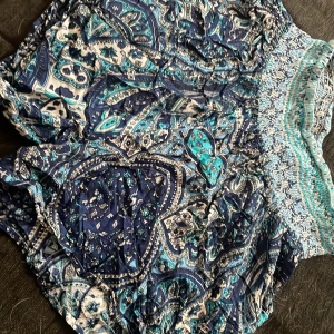 Blå mönstrade shorts med smockad midja - Säljer ett par blå shorts med vitt och turkost paisleymönster. De har en bred, smockad midja som är elastisk och ger en bekväm passform. Lätta och luftiga, perfekta för varma dagar.