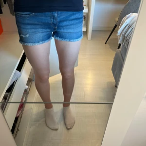 Lågmidjade jeansshorts  - Säljer ett par blå Lågmidjade jeansshorts med fransig kant och dubbla knappar i midjan. Shortsen har klassiska fickor fram och bak samt en liten sliten detalj framtill. Perfekta för varma dagar och enkel att matcha med olika toppar. Jeansshortsen är från H&M och är i storlek 164, men skulle säga att de passar XXS,XS och S. 
