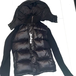 Sixties Cardigan - Säljer en svart pufferjacka från This is Sixtus i storlek XS. Jackan har stickade ärmar och en avtagbar huva. Dragkedja framtill och vadderad kropp för extra värme. Perfekt för kyliga dagar.