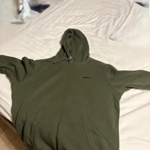Olivgrön hoodie från Nike - Säljer en olivgrön hoodie från Nike med klassisk logga på bröstet. Tröjan har huva med dragsko och en stor magficka framtill. Perfekt för en avslappnad stil.