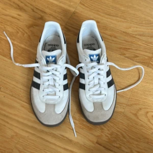 Adidas Samba sneakers i vitt och svart - Klassiska Adidas Samba sneakers i vitt skinn med svarta ränder och grå mockadetaljer vid tån. Använda endast 1 gång då dottern gillar för stora skor och dessa va alltså för små för hennes tycke. Storlek 35 men passar en storlek 36. Innermått 22,5 cm.