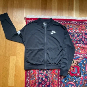 Träningsjacka från Nike - Säljer en svart träningsjacka från Nike med dragkedja framtill och vit Nike-logga på bröstet. Jackan har långa ärmar och ribbade muddar. Perfekt för sport eller vardag när du vill ha en stilren look.