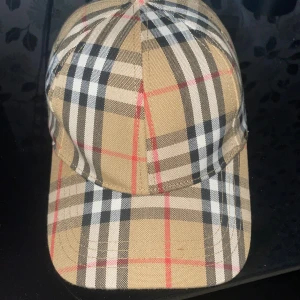 Burberry keps  - Snygg keps i beige med svart, vitt och rött rutmönster. Justerbar rem baktill i metall. Klassisk och tidlös design som passar till många stilar. Tillverkad i bomullsmaterial.
