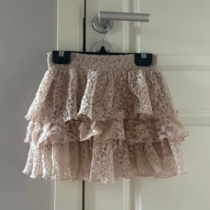 Rosa volangkjol i spets  - Supersöt Rosa kjol med flera lager volanger i spets från Heartquake. Kjolen har dragkedja fram och ett romantiskt, lekfullt uttryck. Perfekt för dig som gillar detaljer och en feminin stil.