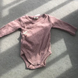 Rosa prickig omlottbody med knytband - Supersöts rosa omlottbody med små prickar och vita knytband på sidan. Långärmad och tillverkad i mjuk bomull, perfekt för mysiga dagar. Tryckknappar i grenen för enkel på- och avklädning.