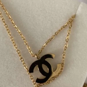 Guldigt halsband med svart Chanel-logo - Säljer ett elegant halsband från Chanel med dubbelkedja i guld och ett hänge i form av den klassiska svarta Chanel-loggan. Perfekt för dig som vill ha en lyxig accessoar med ikonisk design.