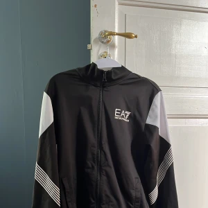 Svart och vit träningsjacka från EA7 Emporio Armani - Snygg svart träningsjacka med vita detaljer och EA7-logga på bröstet. Jackan har dragkedja framtill, ståkrage och långa ärmar med vita och randiga inslag på sidorna. Perfekt för sportiga tillfällen eller en casual look.