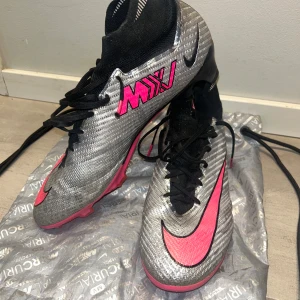 Nike Air Zoom Mercurial Superfly Elite 9 FG - Nike Air Zoom Mercurial Superfly Elite 9 FG. Säljer ett par Nike Mercurial fotbollsskor i silver med rosa detaljer och svart strumpkrage. Skorna har snörning och en platt sula med dobbar för bra grepp på planen. Perfekta för dig som vill sticka ut på fotbollsplanen! Jag köpte fel storlek och skorna är i storlek 46!. För mer bilder/ videos skriv!! Pris kan diskuteras!!