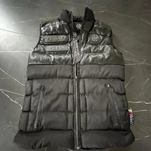  Philipp Plein väst  - Säljer en svart dunväst från Philipp Plein med märkets logga på bröstet och detaljer i skinnimitation upptill. Västen har dragkedja framtill och två fickor med dragkedja. Perfekt för lager-på-lager och har en exklusiv känsla.
