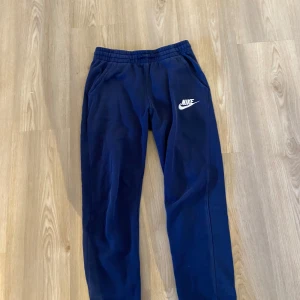 Mörkblå mjukisbyxor från Nike - Mörkblå mjukisbyxor från Nike med vit logga på vänster lår. Byxorna har resår i midjan och sidofickor. Perfekta för en avslappnad stil eller till träning. Byxorna har ett litet hål på låret som är med på bild 3.