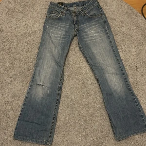 Blå bootcut jeans från Lee - Säljer ett par klassiska blå bootcut jeans från Lee med slitningar och en liten reva på ena benet. Jeansen har fem fickor och Lee-logga bak i midjan. Säljer då de är försmå för mig i midjan! ‼️de är omsydda till låg midjade ‼️skriv om du har frågor!💗