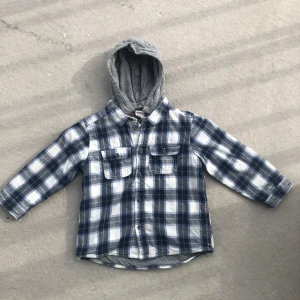 Rutig skjorta med grå luva från H&M - Säljer en blå och vit rutig skjorta från H&M med grå huva i sweatshirtmaterial. Skjortan har långa ärmar, två bröstfickor med knappar och är fodrad med mjukt tyg på insidan. Perfekt för en avslappnad stil.