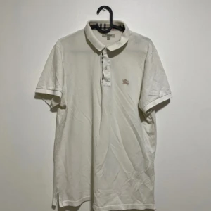 Vit pikétröja från Burberry - Säljer en klassisk vit pikétröja från Burberry med korta ärmar och broderad logga på bröstet. Kragen har Burberrys ikoniska rutiga mönster på insidan och tröjan är tillverkad i bomull. Perfekt för en stilren look.