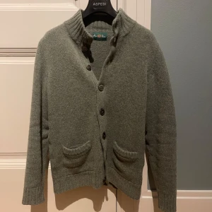 Oliv grön stickad kofta från Alan Paine - Mysig oliv stickad kofta från Alan Paine med hög krage och knäppning framtill. Två fickor framtill och ribbade muddar vid ärmslut och nederkant. Perfekt för lager-på-lager och enkel att matcha!