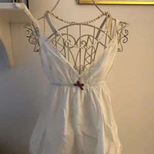 Vit kort topp med spets och rosett - Söt vit blus med v-ringning och spetsdetaljer längs urringningen. Blusen har en liten rosett framtill i midjan och är ärmlös, perfekt för varma dagar.