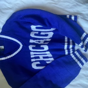 Blå stickad tröja med CHICAGO-tryck - Säljer en blå stickad tröja med vit text 'CHICAGO' framtill. Tröjan har långa ärmar och vita ränder vid halsringning, ärmslut och midja. Perfekt för dig som gillar sportig stil.