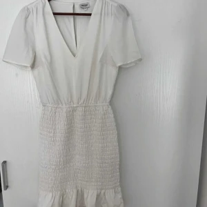 Vit kortklänning med v-ringning - Söt vit klänning med v-ringning och korta ärmar från Bubbelroom. Klänningen har smockad midja och volang nertill, vilket ger en feminin siluett. Perfekt för varma dagar och somriga tillfällen.