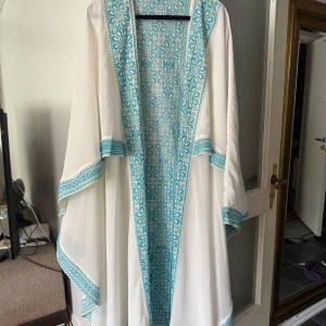 Vit långklänning med turkos broderad kant - Säljer en vit lång kape  med vackra turkosa broderier längs kanterna och ärmarna. Kapen har en lös passform och vida ärmar, perfekt för en luftig och elegant look. Ingen dragkedja eller knappar, bara att slänga på sig. 