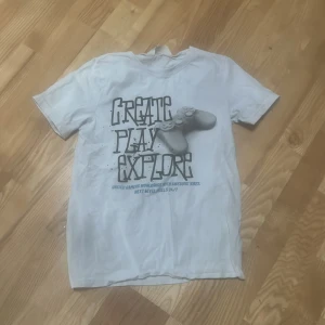 Vit t-shirt med gaming-tryck från H&M - Vit t-shirt från H&M med stort tryck på framsidan som visar en spelkontroll och texten 'Create Play Explore'. Perfekt för dig som gillar gaming och vill ha en avslappnad stil. Använd 1-3 gånger
