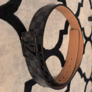 Svart rutigt bälte från Louis Vuitton - Säljer ett stilrent svart bälte från Louis Vuitton med diskret rutigt mönster och svart LV-spänne. Bältet har en slät insida i beige och är tillverkat i skinn. Perfekt accessoar för att lyfta din outfit.