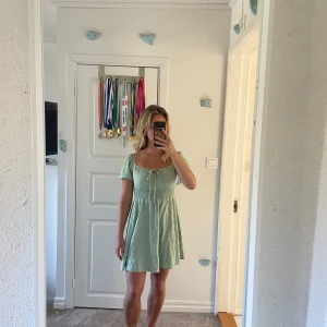 Ljusgrön kortklänning med knyt - Superfin ljusgrön klänning med kort ärm och söt knytning framtill. Klänningen har smock i midjan för en bekväm passform och är tillverkad i ett lätt, luftigt material. 