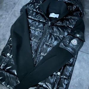 Svart cardigan från moncler  - Snygg svart cardigan från moncler med en glansig framsida och ärmfika med logotyp. I mycket bra skick förutom två knappar i huvan som är dit syda, huvan går att ta av om man tar bort ett par stygn från luvan.