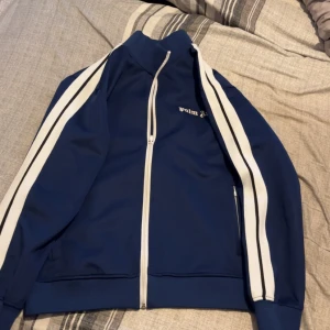 Blå track jacket från Palm Angels - Säljer en blå track jacket från Palm Angels med vita ränder längs ärmarna och vit dragkedja. Jackan har hög krage och logga broderad på bröstet. Perfekt för en sportig och trendig look.