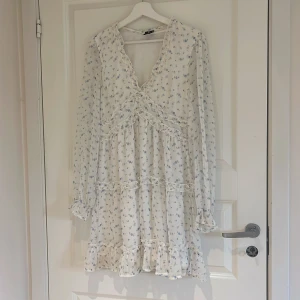 Vit blommig klänning från Vero Moda - Söt vit klänning från Vero Moda med små blå blommor, volangdetaljer och v-ringning. Klänningen har lång ärm och avslutas med en volangkant nertill. Perfekt för vår och sommar! 